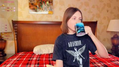 Video of a young woman with shoulder-length brown hair, drinking from a blue and white ceramic mug, wearing a black Metallica t-shirt. She sits on a red and black plaid bedspread in a vintage-style bedroom with floral wallpaper, wooden headboard, and nightstand lamps.