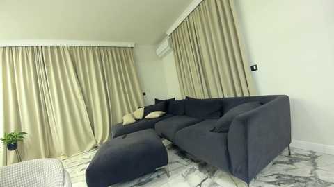 Media: Video of a modern living room with a dark gray sectional sofa, beige curtains, white walls, and a marble-patterned floor.