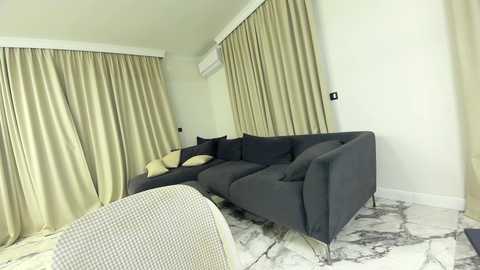 Media: Video of a modern, minimalist living room with a dark gray L-shaped sofa, beige curtains, and marble-patterned flooring.