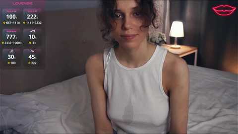 Media: Video of a young, curly-haired woman in a white tank top, sitting on a bed with a grey headboard, beside a bedside lamp, and a TV displaying dating app metrics in the background.