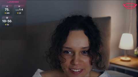 Media: Video of a smiling woman with curly black hair, wearing a white top, in a dimly-lit bedroom with a bedside lamp and a blurred background.