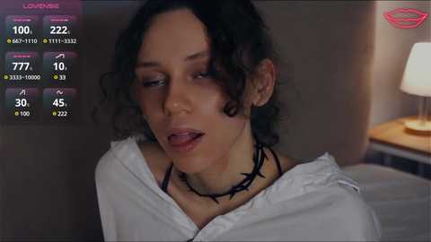 Media: Video of a young woman with curly black hair, wearing a white shirt, black choker, and black makeup, in a dimly lit room with a bedside lamp.