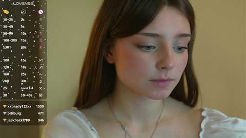 Media: Video of a young woman with light skin, brown hair, and blue eyes, wearing a white blouse, indoors against a beige wall, with a smartphone's camera app interface showing statistics in the background.