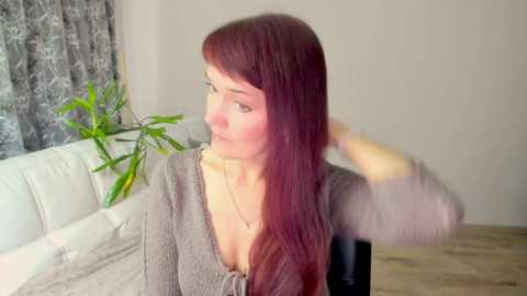 Media: Video of a fair-skinned woman with long, straight auburn hair, wearing a V-neck grey sweater, sitting on a white couch, adjusting her hair. Background includes a potted plant and patterned curtains.