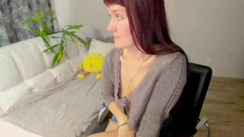 Media: Video of a fair-skinned woman with long, straight, auburn hair, wearing a grey cardigan and beige top, seated on a black office chair in a minimalist room with white sofa, yellow clock, and bamboo plant.