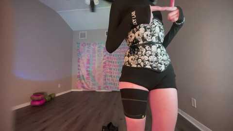 Media: A video of a person with pale skin, wearing a black and white floral corset, black shorts, and thigh-high black stockings, posing in a dimly lit room with a colorful tapestry and scattered workout equipment.