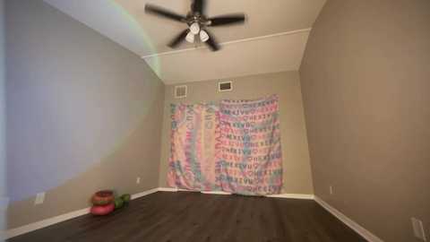 Media: Video of a sparsely furnished, beige-walled room with a ceiling fan, a colorful tie-dye curtain, and a pile of toys on the dark wooden floor.