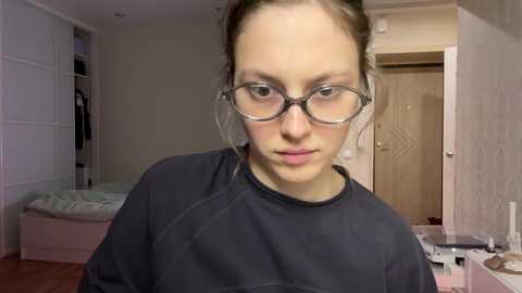 Media: A video of a young woman with light skin, wearing glasses and a black robe, standing in a cluttered bedroom with white walls and a closed door.