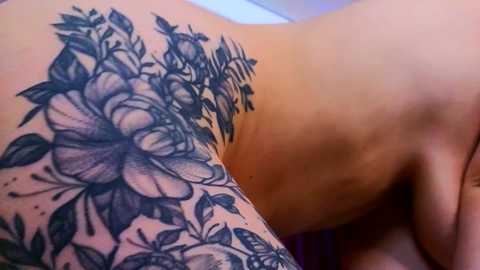 Media: Video of a close-up of a person's shoulder and back, showing intricate black and gray tattoo of large, detailed hibiscus flowers and leaves, with a hand resting on the skin.