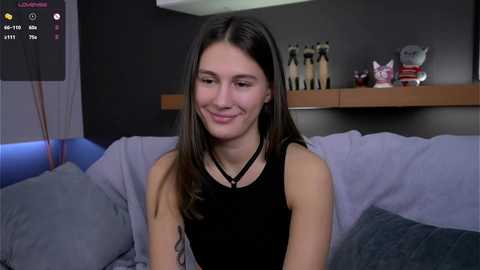 Media: Video of a young woman with long brown hair, fair skin, and a slender build, smiling in a black top, sitting on a light blue couch in a modern living room with dark walls and wooden shelves.