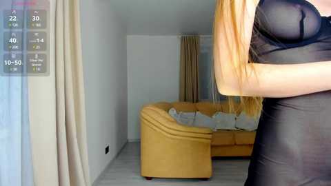 Media: Video of a woman in a sheer black dress standing in a modern living room with beige curtains, a yellow couch, and a TV screen displaying a virtual reality interface.