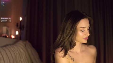 Media: Video of a topless woman with long brown hair, closed eyes, and a serene expression, in a dimly lit bedroom with a bed and soft, ambient lighting.