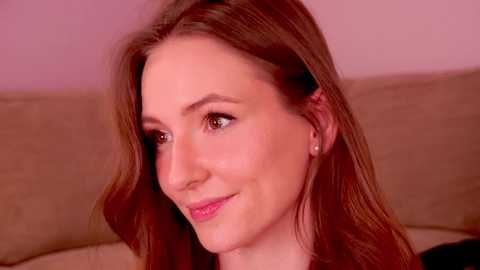 Media: Video of a Caucasian woman with long, wavy, light brown hair, fair skin, and a slight smile, wearing a light-colored top, seated against a beige couch in a softly lit room.