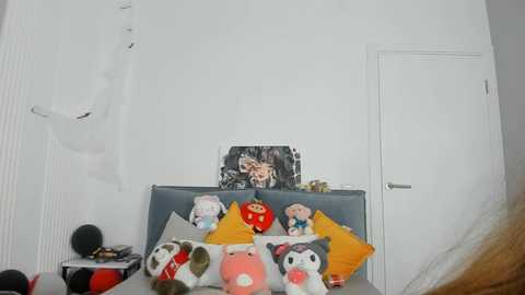 Media: Video of a minimalist room with a white wall and closed door. A grey upholstered headboard holds various plush toys, including Hello Kitty, a red fox, and a pink pig. A framed photo of a man's face hangs above.