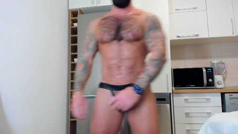 Media: Video of a muscular, tattooed man wearing only black briefs in a modern kitchen, with white cabinets, microwave, and counter.