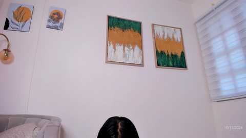 Media: Video of a white wall with two abstract paintings and a lamp, featuring a person with long black hair sitting in a beige couch.