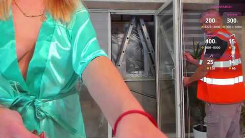 Media: Video: A blonde woman in a low-cut, shiny turquoise blouse, partially revealing her ample cleavage, approaches a glass door. A man in an orange safety vest and helmet stands behind the door.