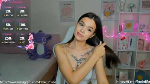 Media: A video of a young woman with long black hair, wearing a sleeveless white top, standing in a pink-lit bedroom. She has a tattoo on her chest. Background includes a purple elephant toy, framed photos, and neon lights.