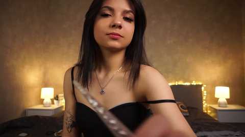 Media: Video of a young, light-skinned woman with long black hair, wearing a black strapless dress, standing in a dimly lit bedroom with warm yellow lighting, a bed, and two nightstands with lamps.