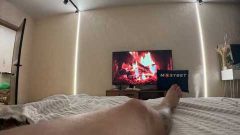 Media: Video of a nude person lying on a bed, with a TV displaying a fireplace scene, illuminated by LED strips on beige walls.