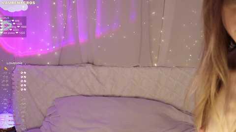 Media: Video of a woman with long blonde hair, partially visible, sitting on a plush white bed with a soft, textured blanket. Background features glowing purple LED lights and a cozy, dimly lit room.