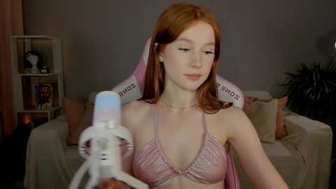 Media: Video of a young, fair-skinned woman with long, straight red hair, wearing a pink bra and pink robe, sitting in a gaming chair, holding a white robot toy, in a cozy, dimly-lit room with shelves and plants.