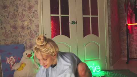 Media: Video of a blonde woman with her hair tied in a bun, wearing a blue sleeveless top, standing next to a white door with frosted glass panels. A plush yellow duck toy and a green neon sign are visible in the background.