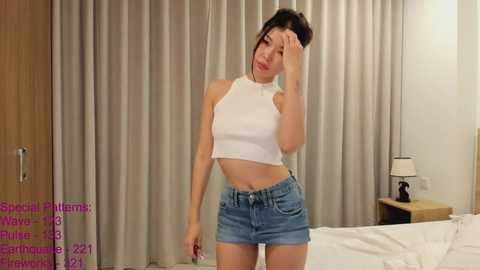 Media: Video of a slender Asian woman with black hair, wearing a white crop top and denim shorts, standing in a modern bedroom with beige curtains.