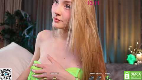 Media: Video of a fair-skinned, slender young woman with long blonde hair, partially covering her breasts with her hands, in a cozy, dimly lit room with a green blanket and plants in the background.