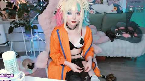 Media: Video of a young woman in cosplay as a character from the anime \"My Hero Academia,\" with vibrant multicolored hair, an orange jacket, black lingerie, and a plush toy. She sits in a cozy room with a cat.