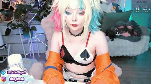 Media: Video of a fair-skinned woman with multicolored hair, wearing a black bra, orange gloves, and a Joker-themed maid outfit, seated in a cozy room with plushies, plants, and a bed.