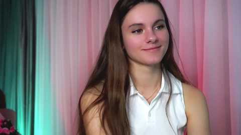 Media: Video of a young, light-skinned woman with long, straight brown hair, wearing a sleeveless white polo shirt, smiling gently, set against a backdrop of pink and green curtains.