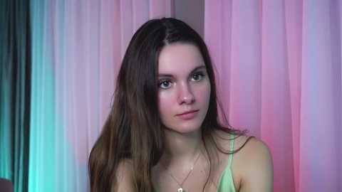 Media: Video of a young Caucasian woman with long brown hair, wearing a light green tank top, standing against pastel-colored curtains. She has fair skin and a neutral expression.