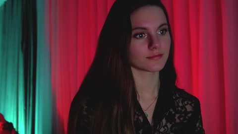 Media: Video of a fair-skinned woman with long black hair, wearing a black lace top, against a red and green draped backdrop.