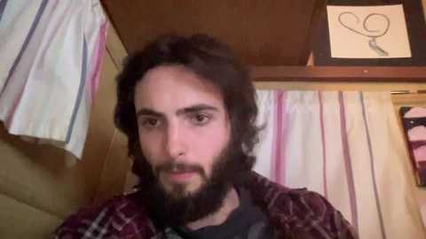 Media: A video of a man with long dark hair and a beard, wearing a plaid shirt, standing in a rustic room with hanging clothes and a wall-mounted drawing.