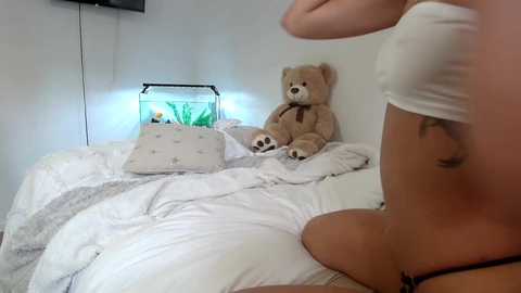 Media: Video of a woman in a white tank top and a teddy bear, lying on a messy white bed with a fish tank and pillows.