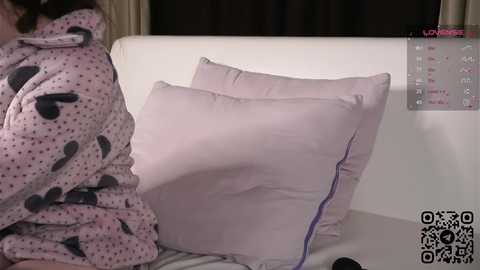 Media: Video of a person in a pink, polka-dotted robe sitting on a bed, with two light purple pillows, a calendar, and a QR code visible in the background.