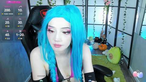 Media: Video of a young woman with bright blue hair, wearing black elbow-length gloves, sitting in a cozy room with a green beanbag, a black chair, and a hanging plant.