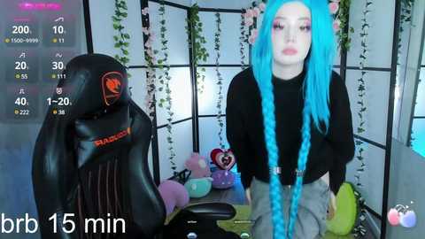 Media: A video of a woman with vibrant blue braided hair, wearing a black turtleneck and gray pants, sitting at a gaming desk. The room is decorated with pastel-colored toys and a black cat plush.