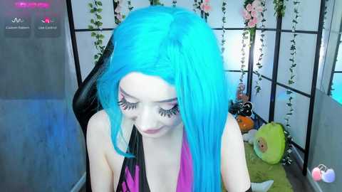 Media: Video of a pale-skinned woman with vibrant blue hair and long eyelashes, wearing a black and pink top, sitting in a room with white walls, pink flowers, and a stuffed toy.