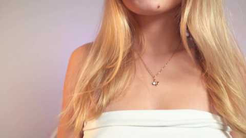 Media: Video of a young woman with long, straight blonde hair, fair skin, and a strapless white top. She wears a delicate gold necklace with a small pendant. The background is a soft, neutral gradient.