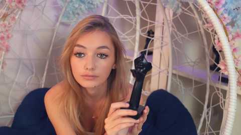 Media: Video of a blonde, fair-skinned woman with blue eyes, lying on a dark surface, using a black hair straightener on her hair. Background features a white, decorative chair with floral accents and a shelving unit.