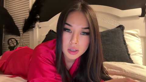 Media: Video of an Asian woman with long, straight, dark hair and light skin, wearing a bright pink satin robe, lying on a bed with white pillows and a black fan in the background.