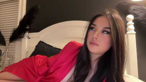 Media: Video of a young woman with long, straight brown hair, wearing a pink robe, reclining on a white bed with black pillows. She gazes thoughtfully into the distance, surrounded by black feather decor.