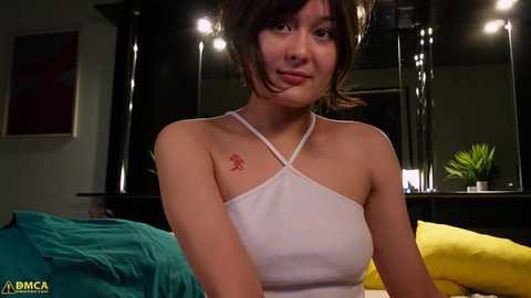 Media: Video of a young Asian woman with short brown hair and a small red tattoo on her left shoulder. She wears a white halter top and sits on a bed with green and yellow bedding.