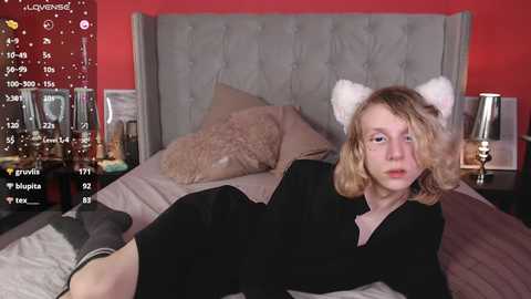 Media: A video shows a young woman with light skin and shoulder-length blonde hair, wearing a black dress and cat ears, lying on a bed with a tufted grey headboard and red walls.