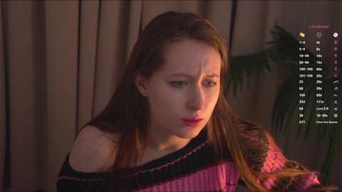 Media: Video of a young Caucasian woman with long, straight brown hair, wearing a pink and black striped sweater, looking concerned. Background features a green plant and beige curtains.