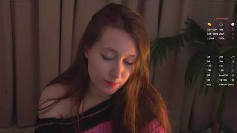 Media: Video of a young woman with long, straight brown hair, wearing a pink and purple striped sweater, indoors with dim lighting, eyes closed, looking down, background includes a potted plant.