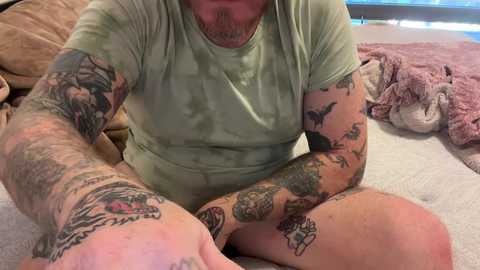 Media: Video of a tattooed, bearded man in a light green shirt, sitting cross-legged on a beige blanket, with plush toys and a teddy bear in the background.