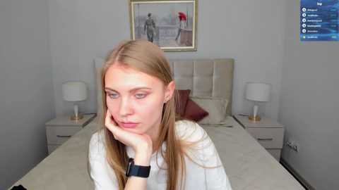 Media: Video of a young Caucasian woman with straight, long blonde hair, blue eyes, and fair skin, resting her chin on her hand in a white blouse. She sits on a neatly made bed with beige linens and pillows. The room has a minimalist decor with a framed picture on the wall.